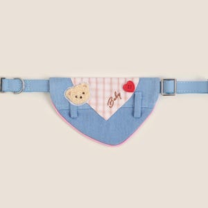 May include: A blue and pink dog bandana with a bear applique, a red heart button, and the text "Baby" embroidered on it. The bandana is attached to a blue adjustable collar with silver buckles.