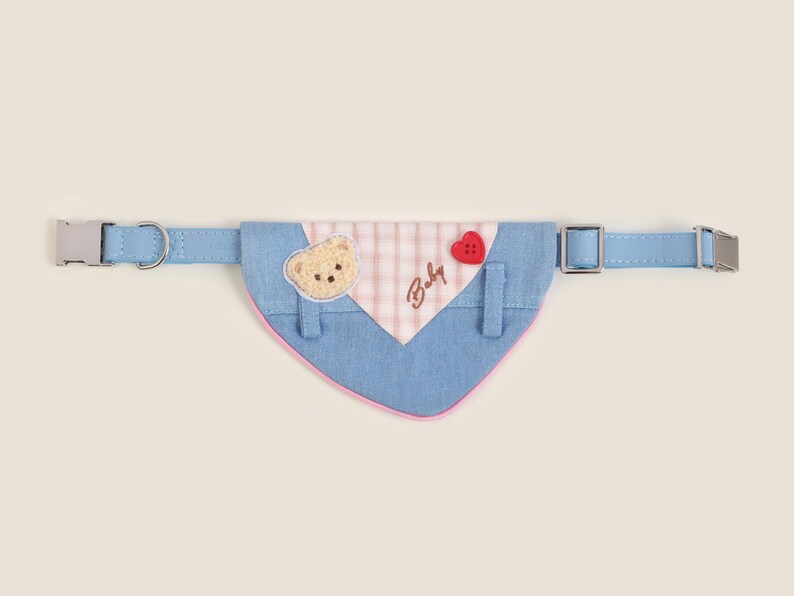 May include: A blue and pink dog bandana with a bear applique, a red heart button, and the text "Baby" embroidered on it. The bandana is attached to a blue adjustable collar with silver buckles.