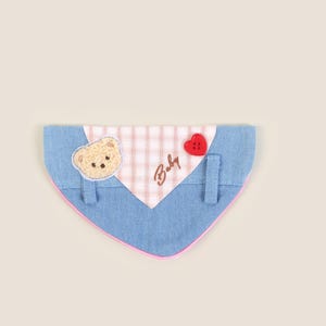 May include: A blue denim dog bandana with a pink and white checkered patch. The patch has the word "Baby" embroidered on it and a small brown bear applique. A red heart-shaped button is attached to the bandana.