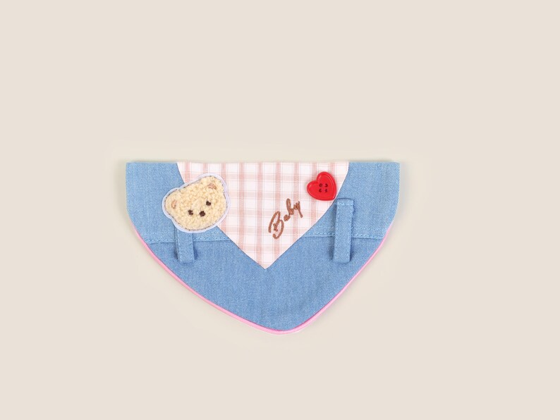 May include: A blue denim dog bandana with a pink and white checkered patch. The patch has the word "Baby" embroidered on it and a small brown bear applique. A red heart-shaped button is attached to the bandana.