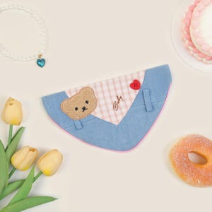 May include: A light blue and pink dog bandana with a brown bear applique, a red heart, and the word "Baby" embroidered on it.