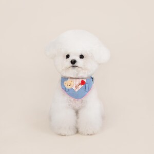 May include: A white dog wearing a blue denim bandana with a pink trim and a small bear applique. The bandana has the word "Baby" embroidered on it.
