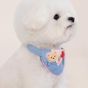 May include: A white dog wearing a blue denim bandana with a pink trim. The bandana has a small embroidered bear and the word "Boh" on it.