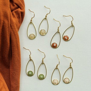May include: Six pairs of gold teardrop-shaped earrings with different colored resin cabochons. The cabochons are in shades of green, peach, and brown.