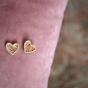 May include: Two small gold heart-shaped ornaments with a floral pattern inside. The ornaments are on a pink velvet surface.