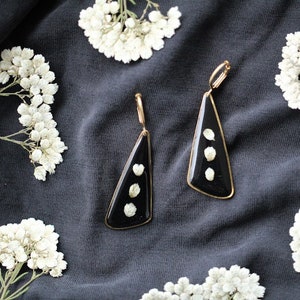 May include: A pair of black triangle-shaped earrings with gold accents. Each earring features three small white flowers encased in resin.