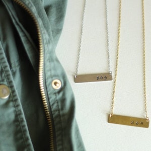 May include: Two gold bar necklaces with the number 365 engraved on them. The necklaces are on a white surface with a green jacket in the background.