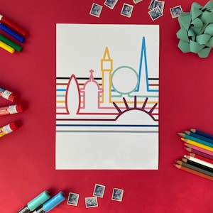 Puede incluir: Una impresión blanca con un diseño de horizonte colorido, que incluye un sol, edificios y formas geométricas. La imagen está rodeada de materiales de arte como lápices de colores, tubos de pintura y rotuladores, todo sobre un fondo rojo.