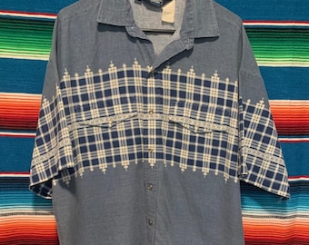 May include: A short-sleeved denim shirt with a blue and white plaid pattern across the chest and sleeves. The shirt has a button-down front and two chest pockets. The shirt is hanging on a black hanger.