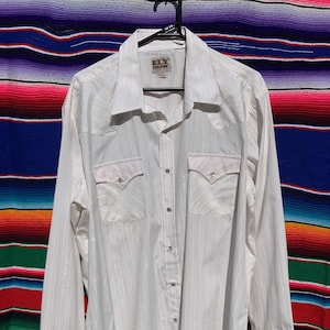 May include: A white long-sleeved shirt with a vertical stripe design. It has two chest pockets and a pointed collar. The shirt is suspended on a black hanger. The brand label "ELY Cattleman" is visible.