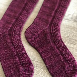May include: A pair of knitted socks in a deep burgundy colour with a cable knit pattern. The socks are made with a soft, textured yarn.