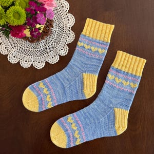 May include: A pair of knitted socks with a blue base, yellow cuffs, heels, and toes. The socks feature a pink heart pattern and a yellow stripe near the cuff. The socks are displayed on a brown surface.