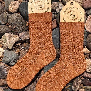 Pode incluir: Um par de meias de lã castanhas com um padrão texturizado. As meias são exibidas em um fundo de pedras e folhas. As meias têm uma etiqueta anexada que diz "The Crazy Sock Lady" com o nome "Kay" em um círculo.