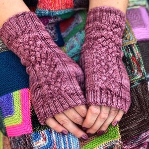 Puede incluir: Un par de guantes sin dedos de punto rosa con un patrón de punto de cable. Los guantes se muestran sobre un fondo colorido de cuadrados de punto.