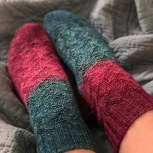 May include: A pair of hand-knitted socks with a gradient color pattern. The socks are green and red with a speckled yarn. The socks are worn on a person's feet.