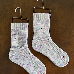 May include: Two hand-knitted socks displayed on metal hangers. The socks are mainly light grey with flecks of pink, purple, and green. The ribbed cuffs and heels are visible, showcasing the detailed craftsmanship of the knitwear.