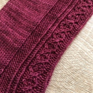May include: Close-up of a knitted fabric with a cable pattern. The fabric is a deep burgundy colour.