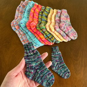 Può includere: Una collezione di piccoli calzini lavorati a maglia a mano in varie colorazioni variegate, tra cui sfumature di blu, arancione, rosa, giallo e verde. I calzini presentano un polsino a coste e una classica costruzione del tallone a ventaglio.