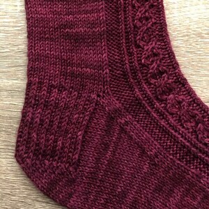 May include: Close-up of a burgundy knitted sock with a cable pattern on the heel and a plain knit on the toe.