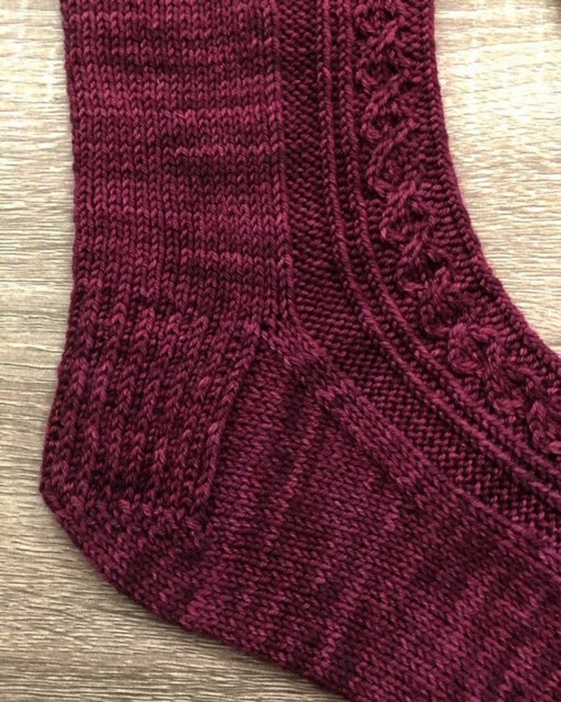 May include: Close-up of a burgundy knitted sock with a cable pattern on the heel and a plain knit on the toe.