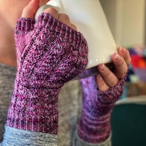 May include: A pair of purple and white fingerless gloves with a textured knit pattern. The gloves are being worn by a person holding a white mug.