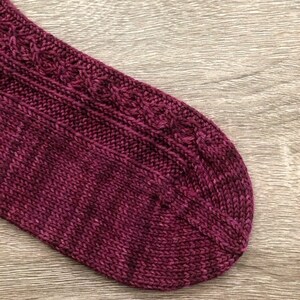 May include: Close-up of a burgundy knitted sock with a cable pattern.