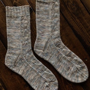 Pode incluir: Um par de meias de tricô em bege e azul com um padrão de cabo. As meias estão colocadas sobre uma superfície de madeira.