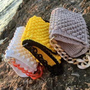 May include: Three crocheted handbags with different colored textured fabric and chain straps. The bags are on a rocky surface.