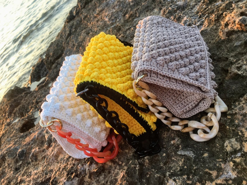 May include: Three crocheted handbags with different colored textured fabric and chain straps. The bags are on a rocky surface.