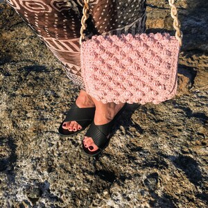 May include: A pink crocheted bag with a chunky chain strap. The bag is worn over the shoulder of a person wearing black sandals.