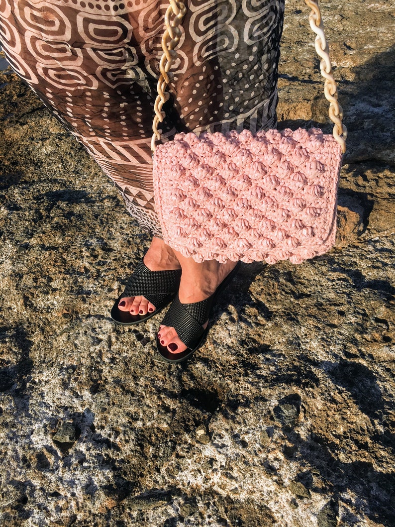 May include: A pink crocheted bag with a chunky chain strap. The bag is worn over the shoulder of a person wearing black sandals.