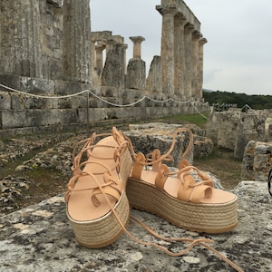 May include: A pair of tan leather platform sandals with lace-up fastenings. The sandals are photographed in front of an ancient stone structure.