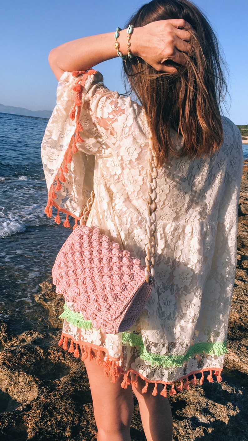 May include: A woman wearing a white crochet dress with pink and green trim and tassels. She is carrying a pink crocheted bag with a white shell chain strap.