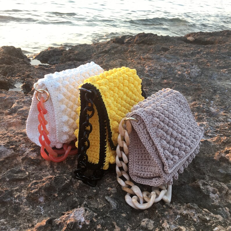 May include: Three crocheted handbags in white, yellow, and beige with different chain straps. The white bag has a red chain strap, the yellow bag has a black chain strap, and the beige bag has a light brown chain strap.