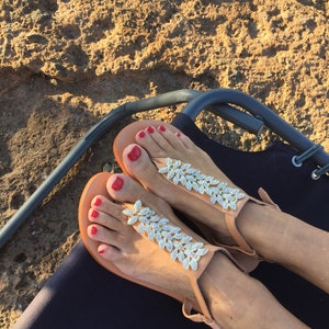 May include: A pair of tan leather sandals with gold leaf embellishments and rhinestones. The sandals have a toe thong and ankle strap. The toes are painted red.