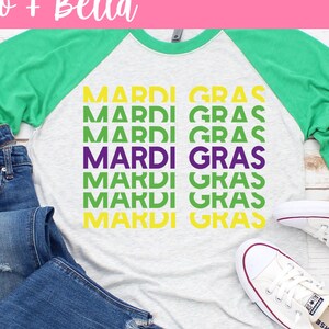 May include: A white, grey, and green long-sleeved shirt with the text "Mardi Gras" repeated multiple times in a stacked design. The shirt is styled with blue jeans and white sneakers.