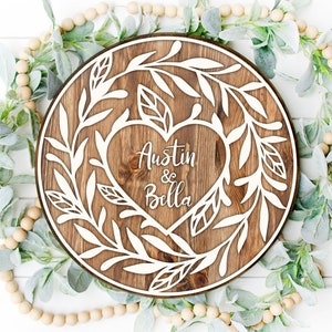 May include: A round wooden sign with a white heart in the center. The heart has the names "Austin & Bella" written inside. The heart is surrounded by white leafy branches.