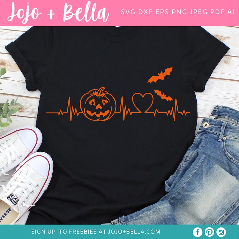 May include: A black t-shirt featuring an orange pumpkin, heart, and bats graphic. The graphic is a heartbeat with a pumpkin, heart, and bats. The t-shirt is laid out on a wooden surface with a pair of white trainers and blue jeans.
