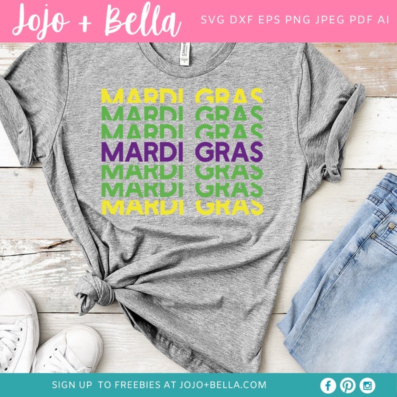 May include: A gray t-shirt with the text "MARDI GRAS" repeated in green, purple, and yellow. The shirt is laid out on a wooden surface with a pair of white sneakers and a pair of blue jeans.
