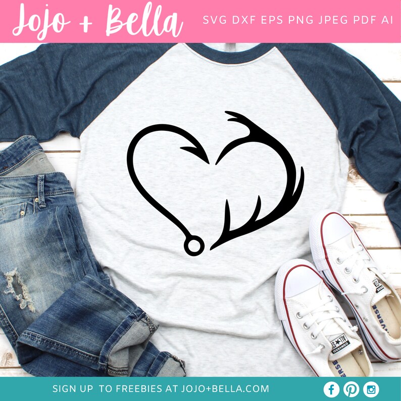 May include: A white long-sleeved shirt with a blue sleeve, featuring a black silhouette of a heart formed by a fishing hook and an antler. The shirt is laid out on a wooden surface with a pair of white sneakers and blue jeans.