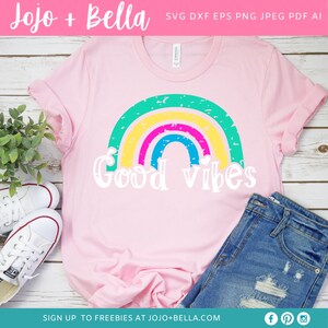Op de afbeelding: Een roze T-shirt met een regenboogontwerp en de tekst "Good vibes" in wit. De regenboog is gemaakt van groene, gele, roze, blauwe en gele strepen.