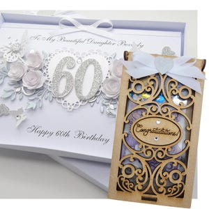 May include: A white 60th birthday card with a decorative floral design and the text "Happy 60th Birthday". A wooden money box with an ornate cutout design and a white bow is next to the card.