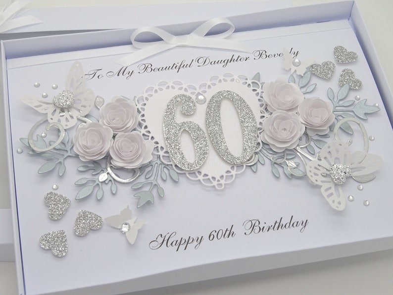 May include: A white birthday card with a decorative design. The card features a silver glitter "60" in the center, surrounded by paper flowers, butterflies, and hearts. The text "Happy 60th Birthday" is written at the bottom.