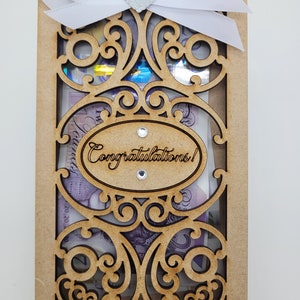 May include: A wooden gift box with an ornate, laser-cut design. The box is adorned with a white ribbon and a silver glitter heart. The words "Congratulations!" are laser-cut on an oval. The box contains money.