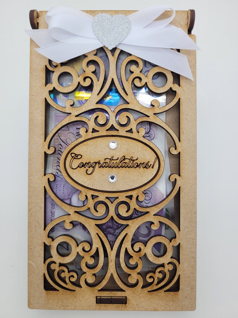 May include: A wooden gift box with an ornate, laser-cut design. The box is adorned with a white ribbon and a silver glitter heart. The words "Congratulations!" are laser-cut on an oval. The box contains money.