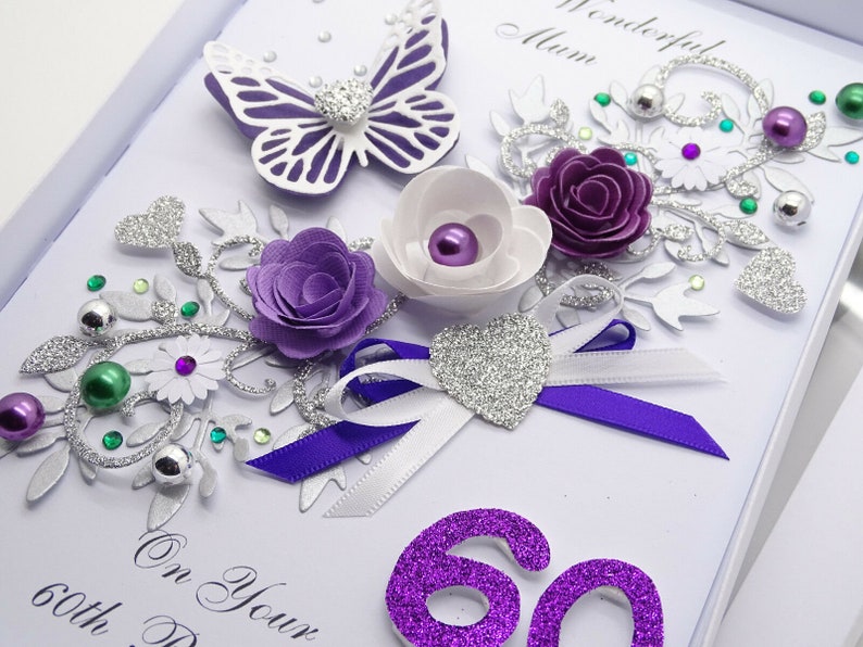 May include: A handmade card with a purple and silver design. The card features a butterfly, flowers, and a ribbon. The number 60 is written in purple glitter. The text "On Your 60th Day" is written in black ink.