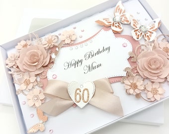 May include: A white box with a pink and gold card inside. The card has a pink and gold floral design with a heart-shaped tag that says "60" on it. The card also has the text "Happy Birthday Mum" on it.