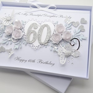 May include: A white 60th birthday card with a decorative design. The card features silver glitter numbers, paper flowers, butterflies, and hearts. The text reads "To My Beautiful Daughter Beverly" and "Happy 60th Birthday".