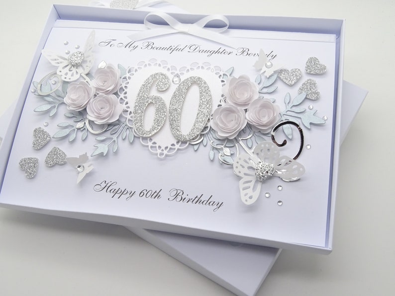 May include: A white 60th birthday card with a decorative design. The card features silver glitter numbers, paper flowers, butterflies, and hearts. The text reads "To My Beautiful Daughter Beverly" and "Happy 60th Birthday".