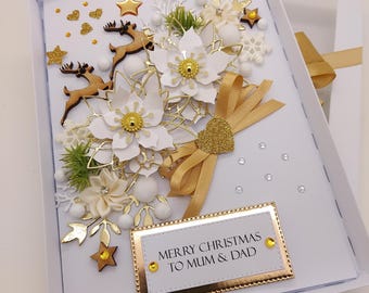 May include: A white rectangular greeting card featuring layered paper poinsettias, gold ribbon, wooden reindeer silhouettes, and glittery stars. A gold-bordered plaque at the bottom reads MERRY CHRISTMAS TO MUM & DAD, accented with small yellow rhinestones.